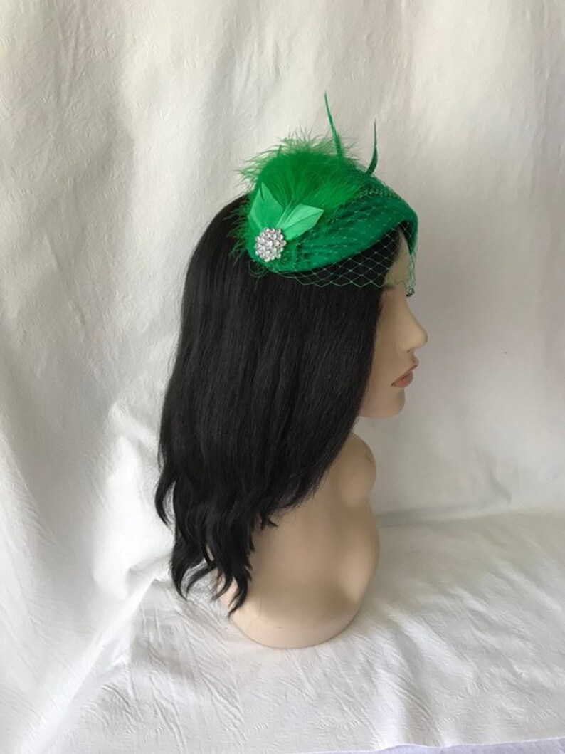 May include: A green hat with a feather and a rhinestone embellishment. The hat is made of a soft, textured fabric and has a veil.