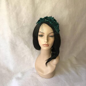 May include: A dark green headpiece with a floral design and a black netting veil. The headpiece is displayed on a mannequin head with black hair and a side braid. The background is a light, textured fabric.