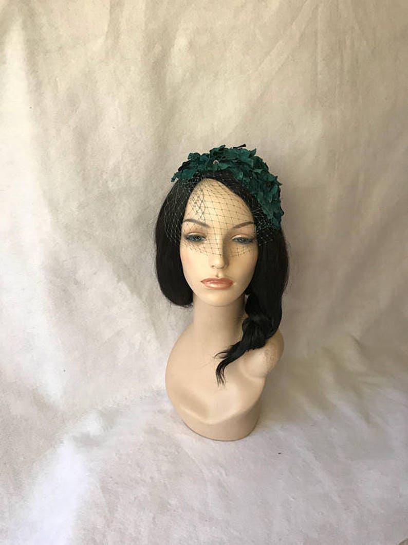 May include: A dark green headpiece with a floral design and a black netting veil. The headpiece is displayed on a mannequin head with black hair and a side braid. The background is a light, textured fabric.