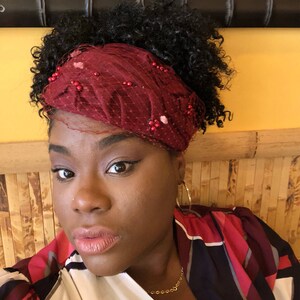 May include: A woman with curly black hair wearing a red headband with beaded embellishments and a red, black and white patterned blouse. She is wearing gold hoop earrings and a gold chain necklace.