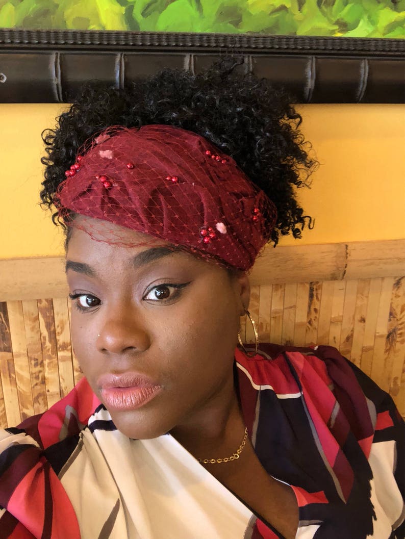 May include: A woman with curly black hair wearing a red headband with beaded embellishments and a red, black and white patterned blouse. She is wearing gold hoop earrings and a gold chain necklace.