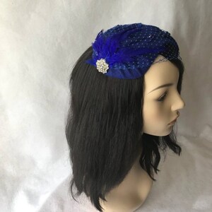 May include: A royal blue fascinator with a small, sparkly brooch and a blue feather accent. The fascinator is made of netting and is perched on a mannequin head with black hair.