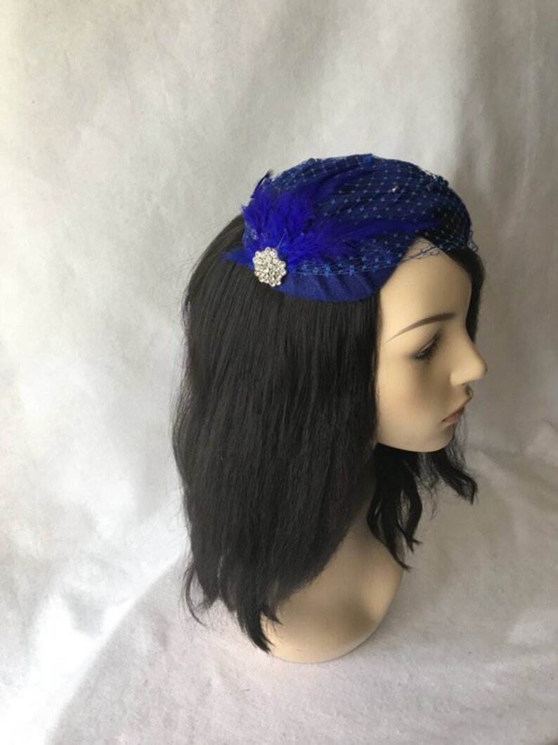 May include: A royal blue fascinator with a small, sparkly brooch and a blue feather accent. The fascinator is made of netting and is perched on a mannequin head with black hair.