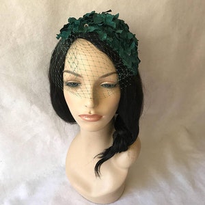 May include: A dark green floral headpiece with a delicate black netting veil. The headpiece is adorned with fabric leaves and a small, sparkling embellishment. The mannequin head has black hair and is set against a white background.