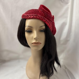 May include: A red, embellished hat with a curved brim and a decorative accent. The hat is covered in sequins and small beads, creating a textured surface. The hat is displayed on a mannequin head with black hair.