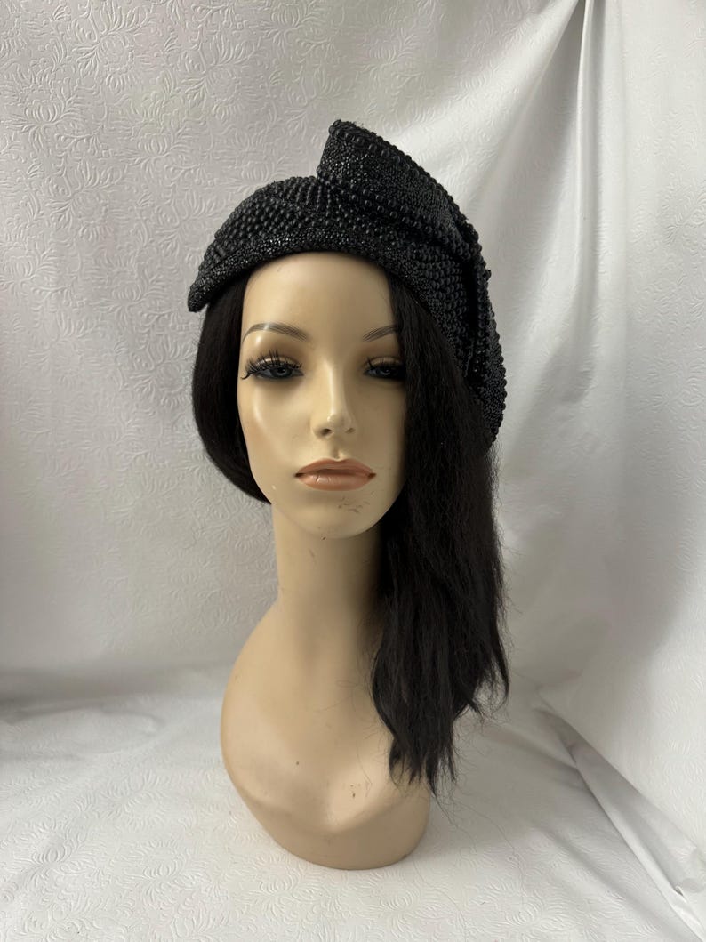 May include: A black beaded hat with a bow on the side. The hat is angled on a mannequin head with black hair.