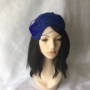 May include: A royal blue felt hat with a netting veil and a blue feather accent. The hat is decorated with small blue beads.