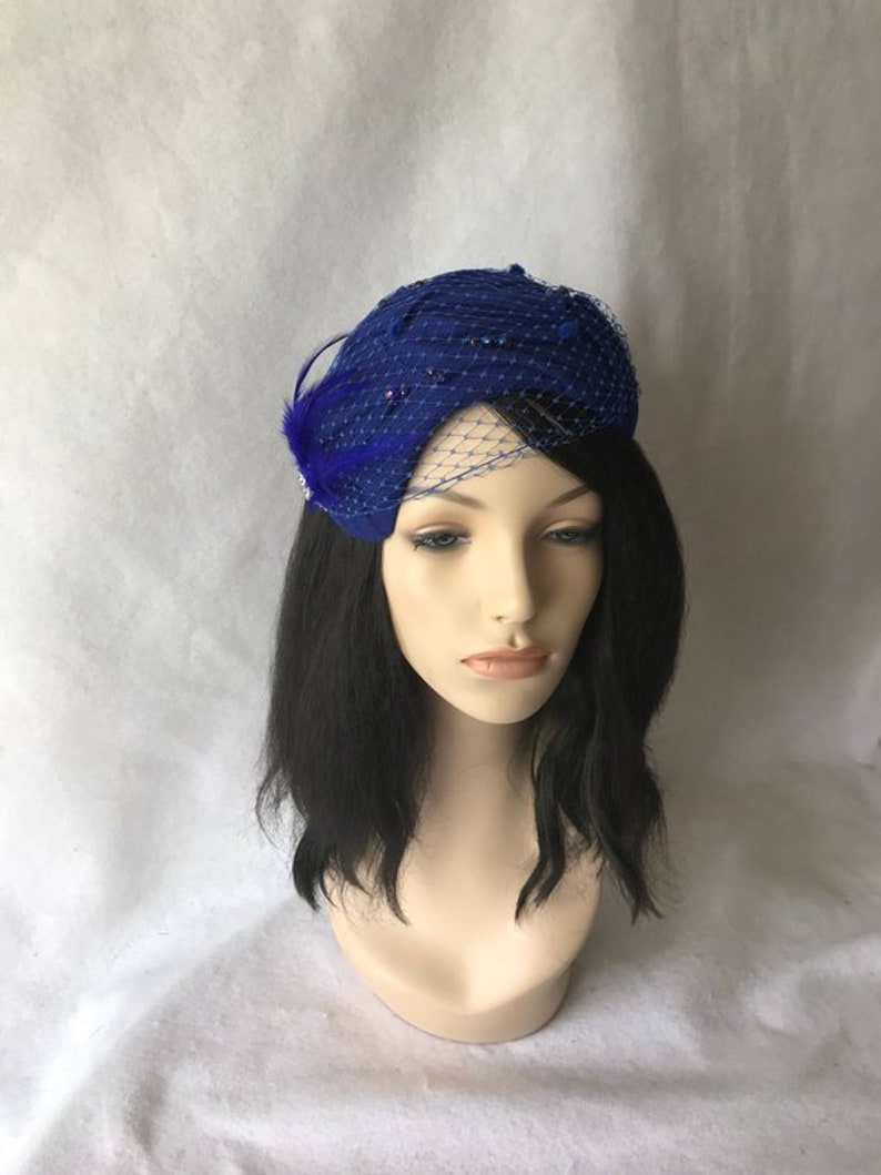 May include: A royal blue felt hat with a netting veil and a blue feather accent. The hat is decorated with small blue beads.