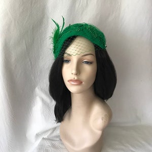 May include: A green felt hat with a beaded design and a small veil. The hat is adorned with a feather accent.