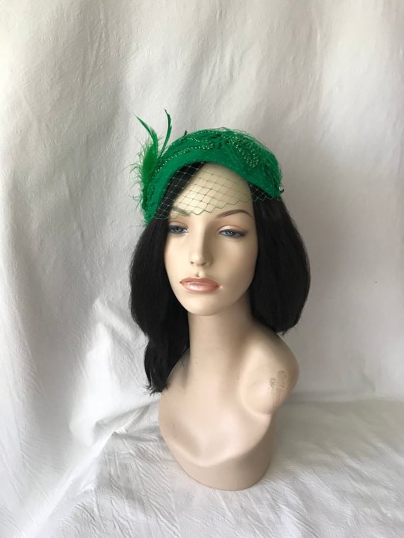 May include: A green felt hat with a beaded design and a small veil. The hat is adorned with a feather accent.