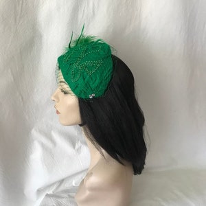 May include: A green felt hat with a beaded design and a feather accent. The hat is worn on a mannequin with black hair.