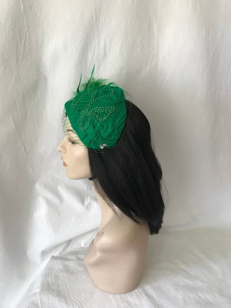 May include: A green felt hat with a beaded design and a feather accent. The hat is worn on a mannequin with black hair.