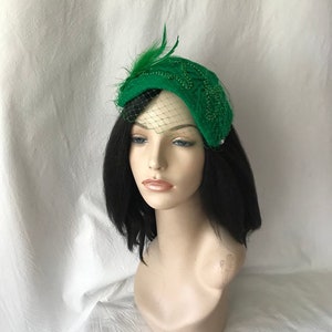 May include: A green felt hat with a beaded design and a feather accent. The hat is adorned with a green tulle veil.