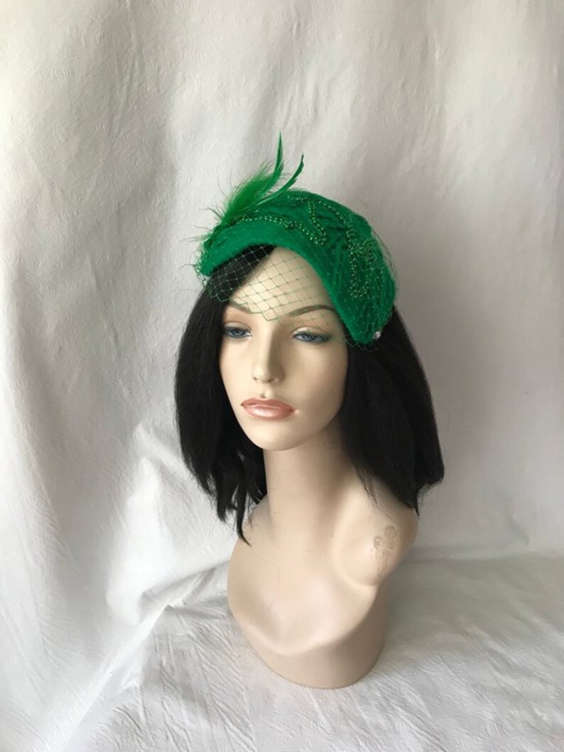 May include: A green felt hat with a beaded design and a feather accent. The hat is adorned with a green tulle veil.