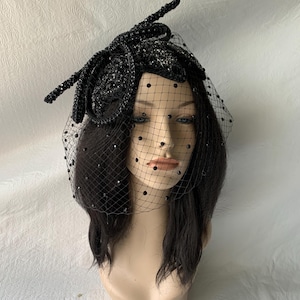 May include: A black headpiece with a decorative design, featuring a veil with small black embellishments. The headpiece is adorned with sparkling accents and a floral-like embellishment. The veil drapes over the face of a mannequin head.