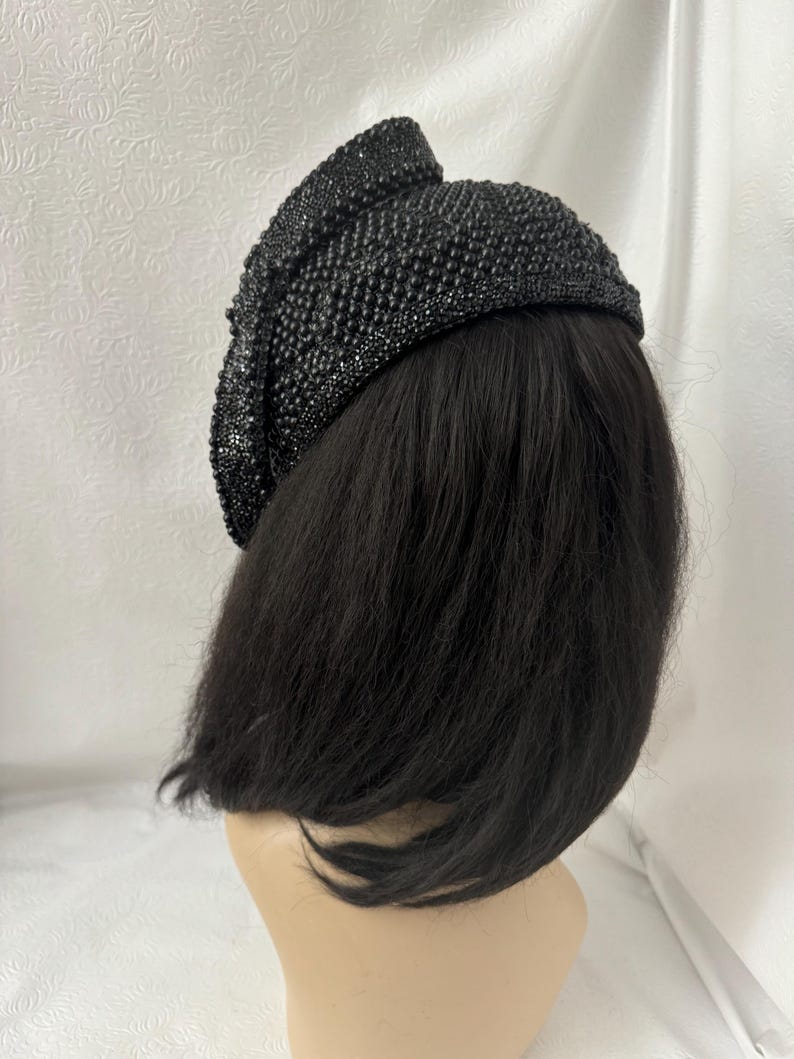 May include: A black beaded hat with a unique design. The hat is made of small, round beads that are sewn together to create a textured surface. The hat is worn on a mannequin with black hair.