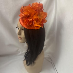 May include: A bright orange flower hat with a round base. The hat is made of fabric and has a cluster of orange fabric flowers on top.