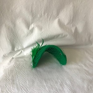 May include: A green hat with a feather and netting. The hat is made of a soft, pliable material and has a curved shape.