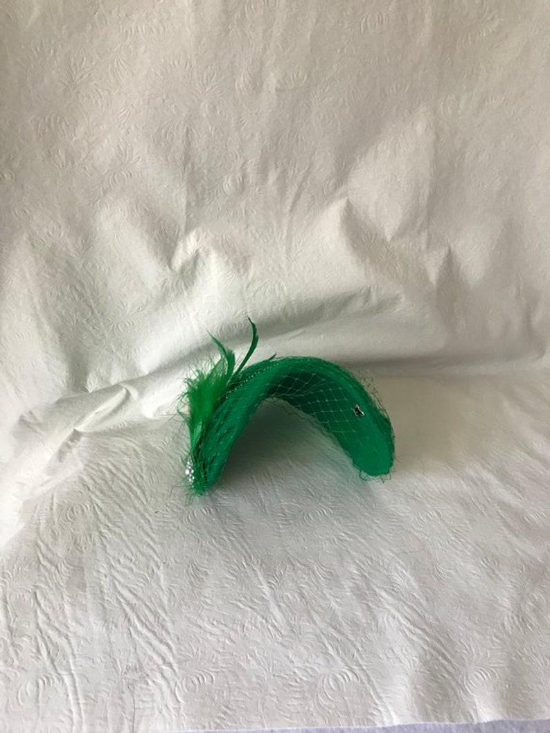 May include: A green hat with a feather and netting. The hat is made of a soft, pliable material and has a curved shape.