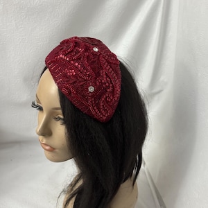 May include: A burgundy sequined headband with a rounded shape. The headband is embellished with small, sparkling sequins and three clear, round rhinestones. The accessory is designed to be worn on the head and adds a touch of glamour.