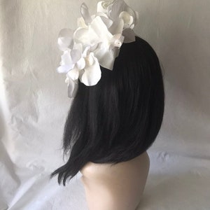 May include: White flower hairpiece with a cluster of large, white fabric flowers. The hairpiece is on a black wig.