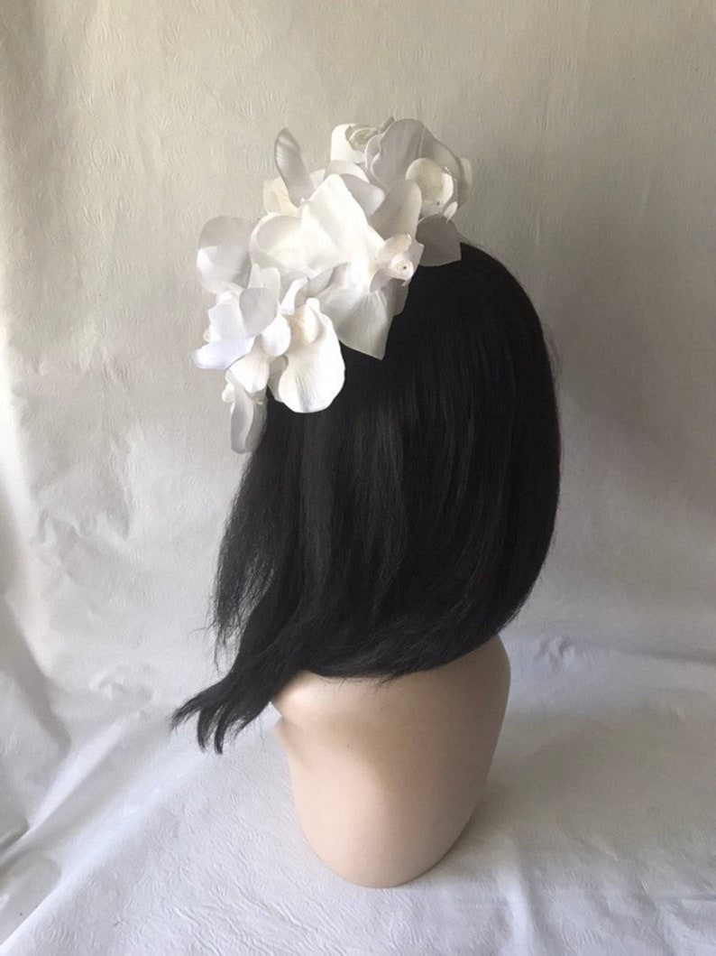 May include: White flower hairpiece with a cluster of large, white fabric flowers. The hairpiece is on a black wig.