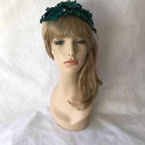 May include: A dark teal floral headpiece with a delicate veil. The headpiece features a cluster of fabric flowers and small pearl accents. The veil is a fine mesh that drapes over the face, creating a vintage aesthetic.