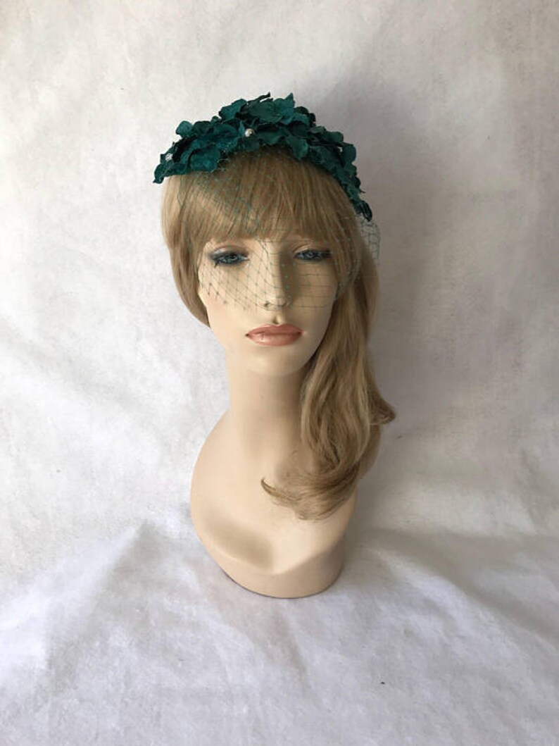 May include: A dark teal floral headpiece with a delicate veil. The headpiece features a cluster of fabric flowers and small pearl accents. The veil is a fine mesh that drapes over the face, creating a vintage aesthetic.