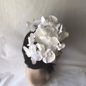 May include: A white flower crown made of fabric orchids. The crown is worn on a mannequin head with black hair.
