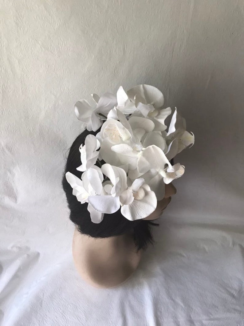 May include: A white flower crown made of fabric orchids. The crown is worn on a mannequin head with black hair.