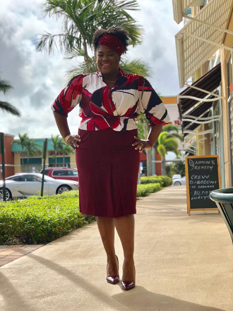 May include: A woman wearing a red, white and black patterned blouse with a burgundy skirt and burgundy heels. She is standing on a pavement in front of a shop with a sign that says "Nature's Remedy, Green Discount, 30 Hours, Blood Analysis".