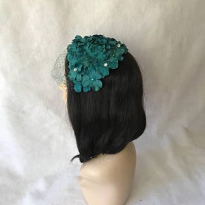 May include: A teal floral headpiece with a delicate veil. The headpiece features clusters of fabric flowers adorned with small, sparkling embellishments. The veil is a fine, sheer mesh. The headpiece is displayed on a mannequin head with black hair.