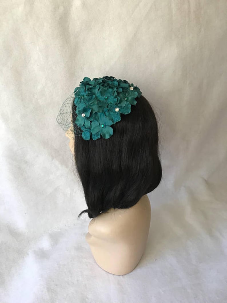 May include: A teal floral headpiece with a delicate veil. The headpiece features clusters of fabric flowers adorned with small, sparkling embellishments. The veil is a fine, sheer mesh. The headpiece is displayed on a mannequin head with black hair.