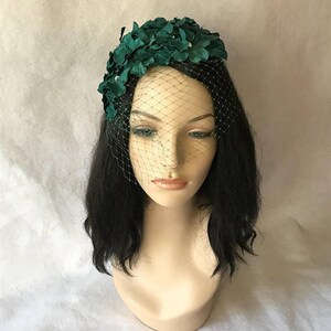 May include: A dark green floral headpiece with a delicate net veil. The headpiece features clusters of fabric flowers and small, sparkling accents. The veil is a fine mesh, adding a touch of vintage elegance. The headpiece is displayed on a mannequin head.