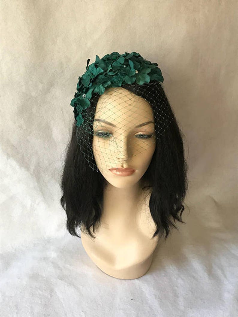 May include: A dark green floral headpiece with a delicate net veil. The headpiece features clusters of fabric flowers and small, sparkling accents. The veil is a fine mesh, adding a touch of vintage elegance. The headpiece is displayed on a mannequin head.