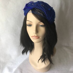 May include: A royal blue felt hat with a netting veil and a feather accent. The hat is adorned with small, dark blue beads.