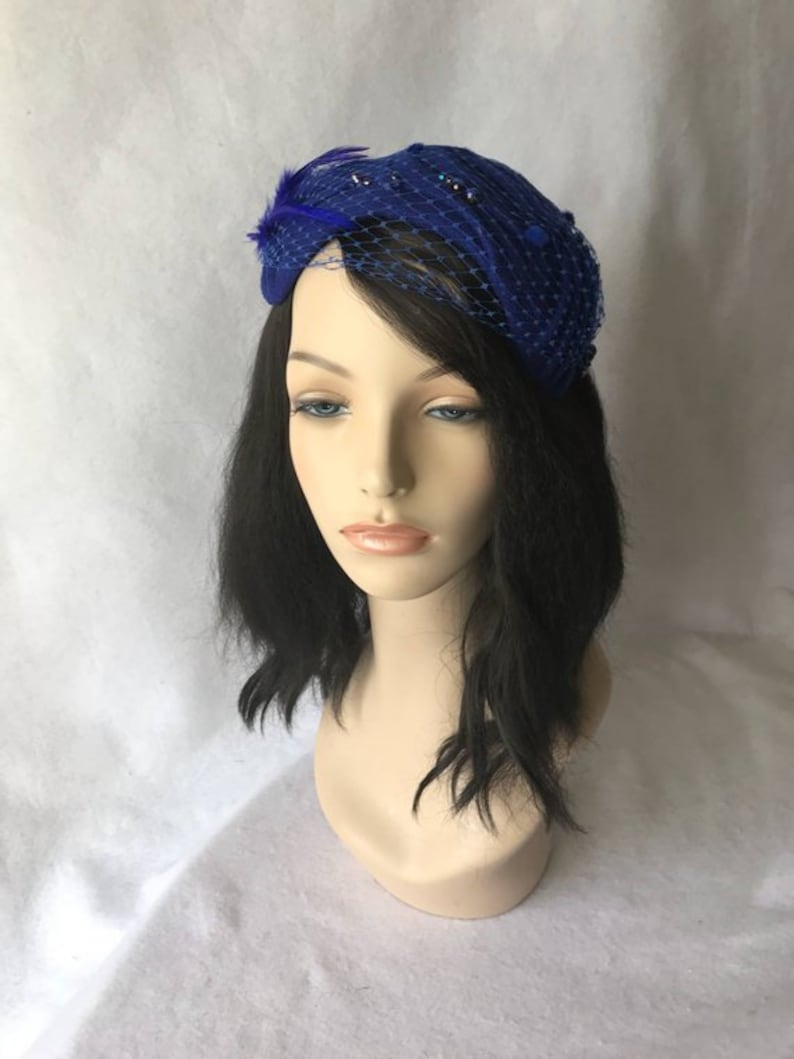 May include: A royal blue felt hat with a netting veil and a feather accent. The hat is adorned with small, dark blue beads.