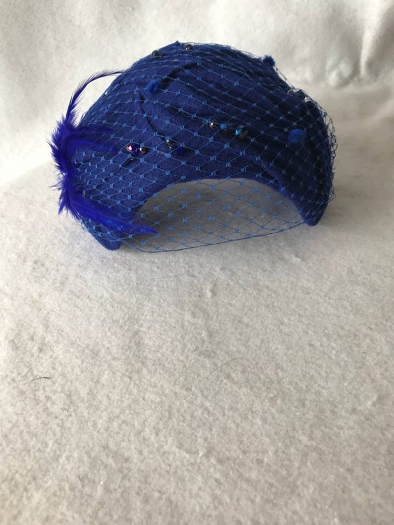 May include: A blue felt hat with a blue veil and blue feather embellishment. The hat is decorated with small, sparkly rhinestones.