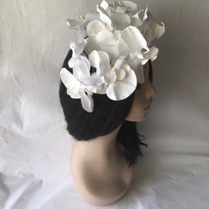 May include: A white flower crown made of artificial orchids. The crown is worn on a mannequin head with black hair.