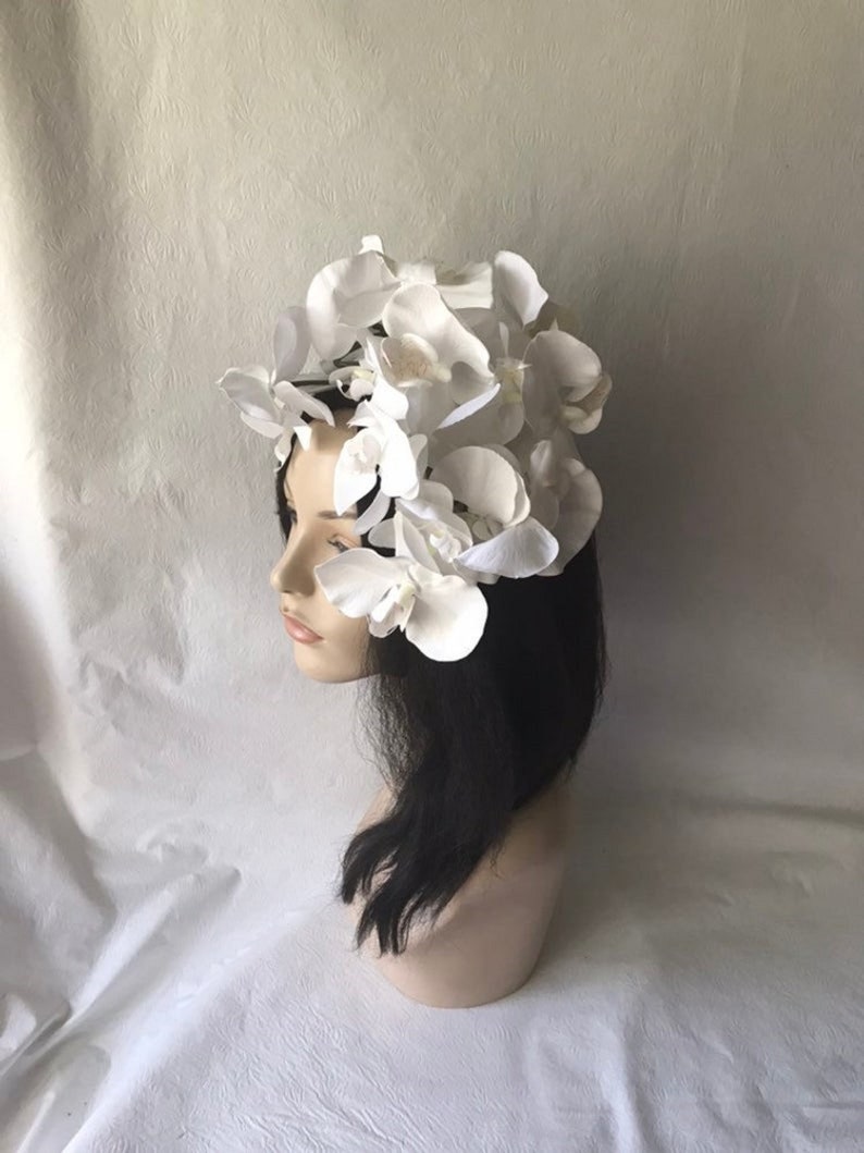 May include: A white flower crown made of artificial orchids. The crown is worn on a mannequin with black hair.