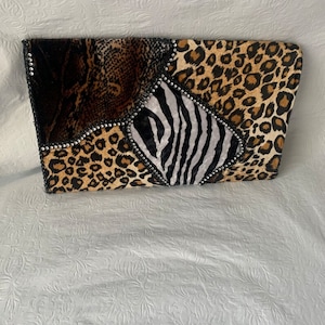 以下が含まれることがあります： ヒョウ柄、シマウマ柄、ヘビ柄の生地を組み合わせたパッチワークデザインの黒いクラッチバッグ。バッグはラインストーンで飾られています。