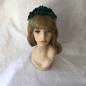 May include: A dark green floral headpiece with a delicate veil. The headpiece is made of fabric and features a cluster of leaves. The veil is a fine mesh that drapes over the face. The headpiece is displayed on a mannequin head.