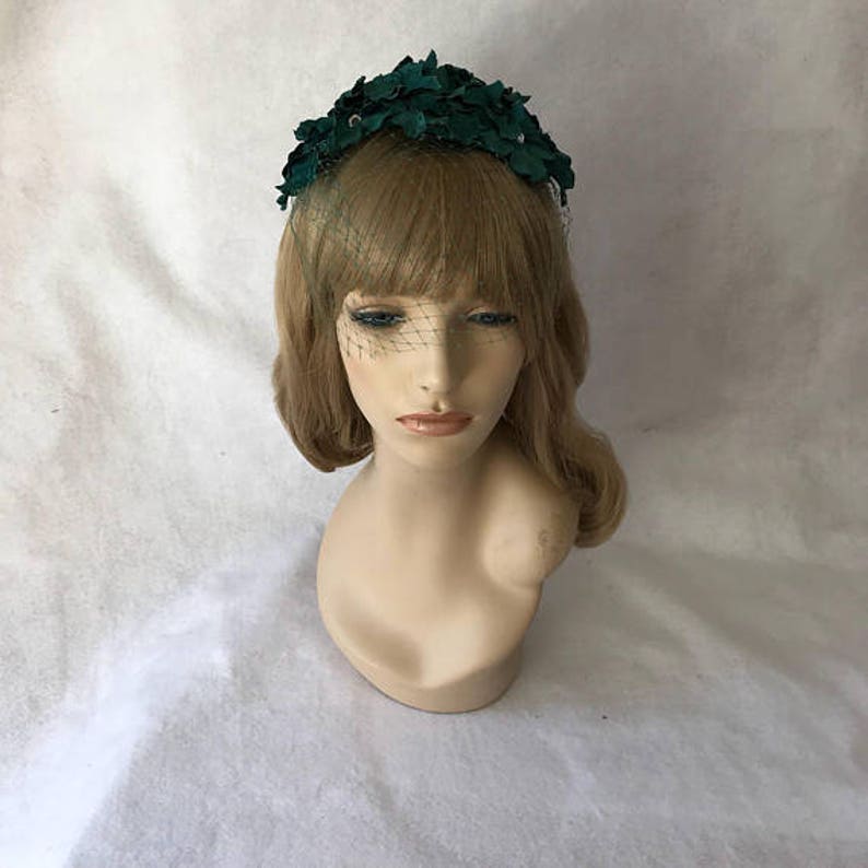 May include: A dark green floral headpiece with a delicate veil. The headpiece is made of fabric and features a cluster of leaves. The veil is a fine mesh that drapes over the face. The headpiece is displayed on a mannequin head.