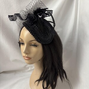 May include: Black headpiece with a decorative design. The headpiece features a beaded base, a net veil, and feather accents. The headpiece is designed to be worn on the head.