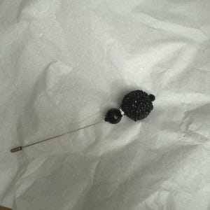May include: A black pin with a round, sparkly black bead at the top. The pin is on a white surface.