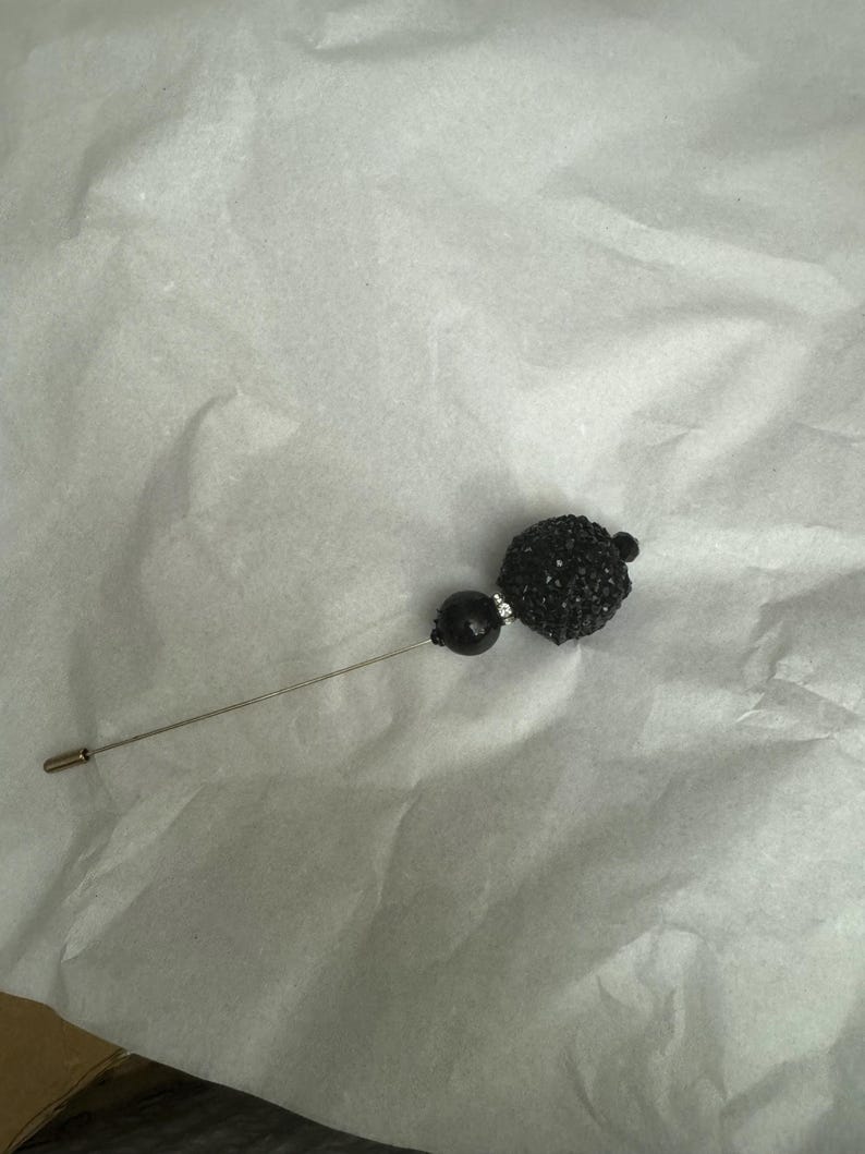 May include: A black pin with a round, sparkly black bead at the top. The pin is on a white surface.