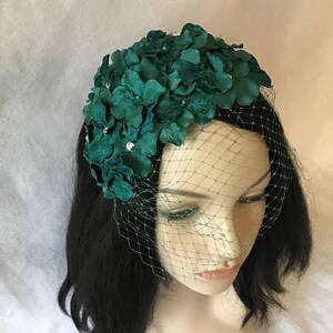 May include: A dark green floral headpiece with a matching veil. The headpiece is adorned with fabric flowers and sparkling embellishments. The veil is a fine mesh, adding a touch of elegance. The headpiece is worn on a mannequin head with dark hair.