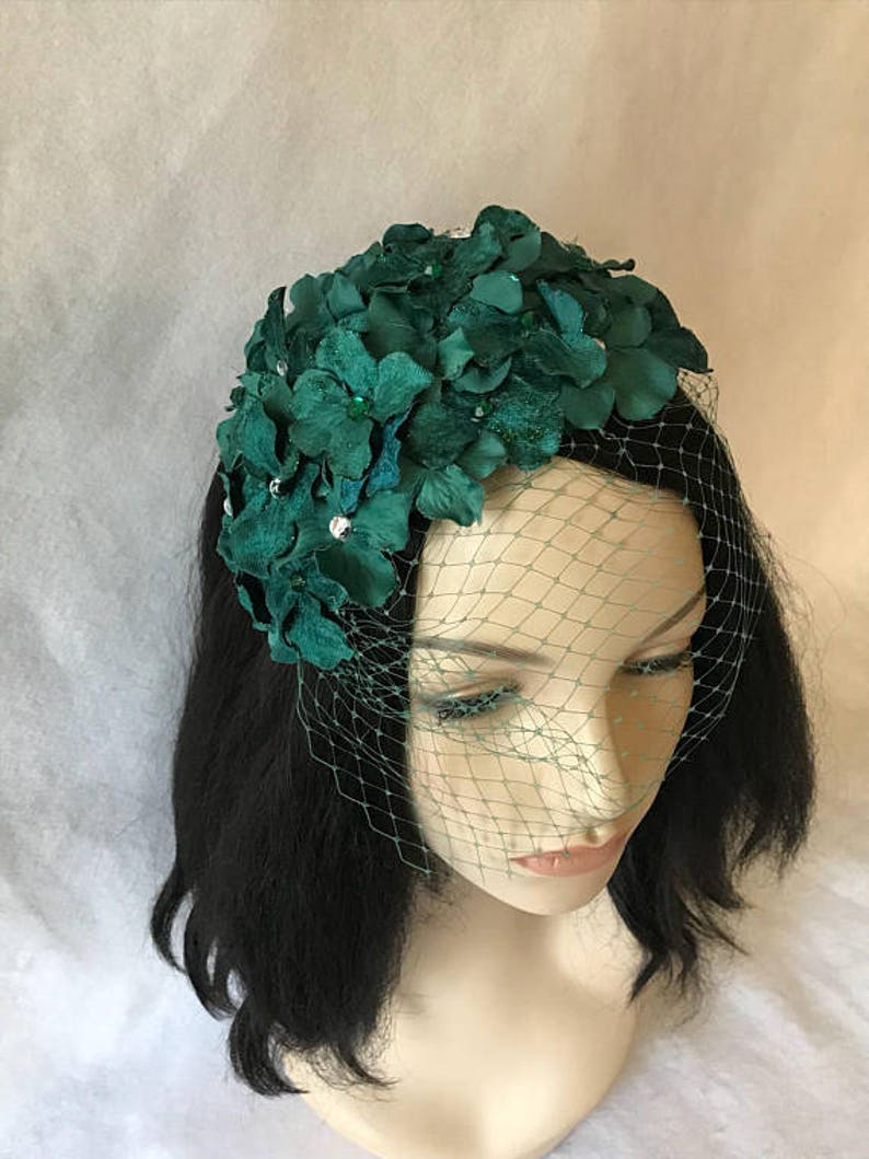 May include: A dark green floral headpiece with a matching veil. The headpiece is adorned with fabric flowers and sparkling embellishments. The veil is a fine mesh, adding a touch of elegance. The headpiece is worn on a mannequin head with dark hair.