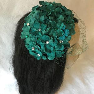 May include: A dark green floral headpiece with a veil. The headpiece features fabric flowers and is adorned with clear and green rhinestones. The veil is a fine, dark mesh.