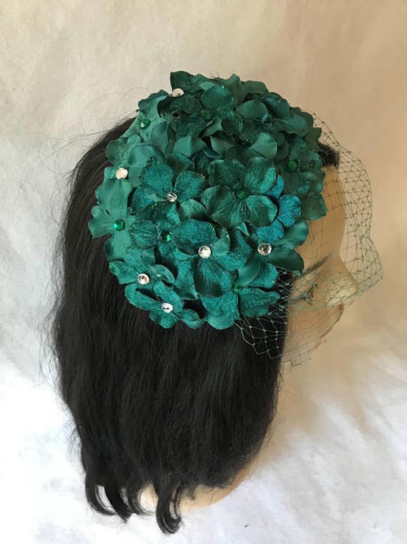 May include: A dark green floral headpiece with a veil. The headpiece features fabric flowers and is adorned with clear and green rhinestones. The veil is a fine, dark mesh.